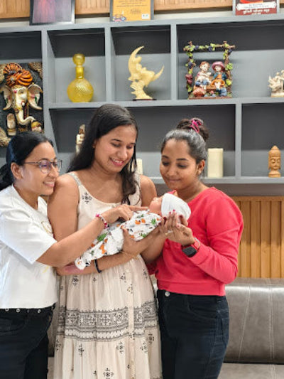 Three women holding a baby in a room with shelves displaying decorative items.